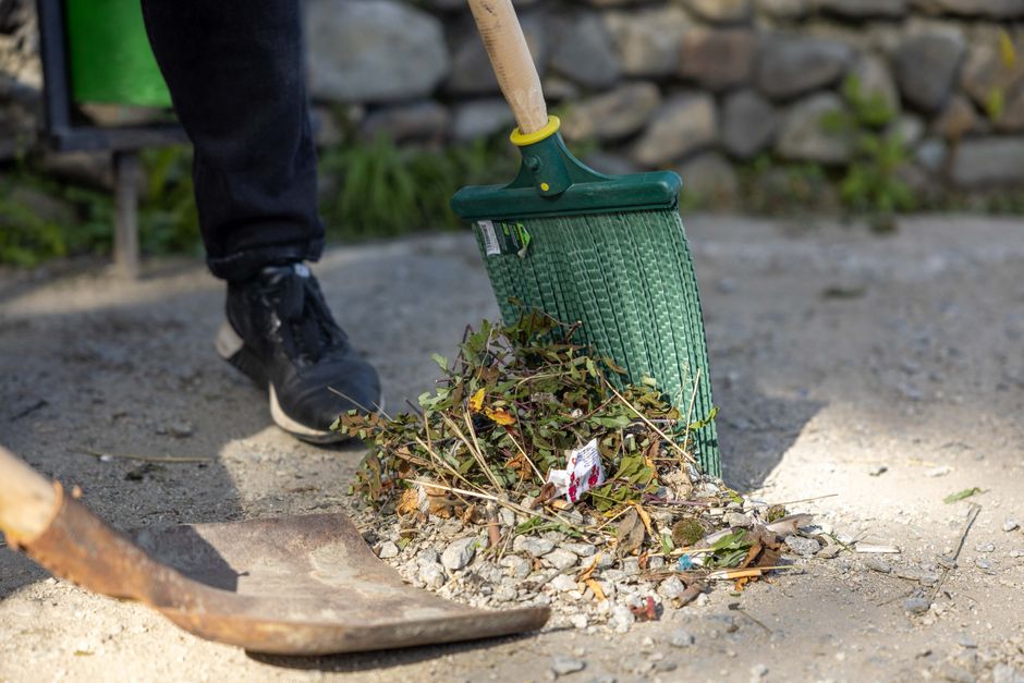 A person wearing black trousers and black sneakers is using a green rubber hand rake to gather fallen leaves, small branches, and garden debris from a gravel pathway in a garden. The rake blade is wide and textured, suitable for collecting loose material. The surrounding area includes a section of compacted soil bordered by a stone wall in the background, with some green plants growing near the base of the wall. The garden appears tidy and well-maintained, with a mix of leaf litter and small twigs on the ground, indicative of ongoing garden maintenance or hedge trimming activities. The lighting suggests it is daytime, with natural light illuminating the scene, emphasizing the textures of the gravel, leaves, and stone wall. This image relates to outdoor garden care, demonstrating the process of clearing debris as part of lawn or hedge trimming, an activity supported by Gardeners Bayswater in line with Westminster Council garden waste rules for Bayswater gardeners.