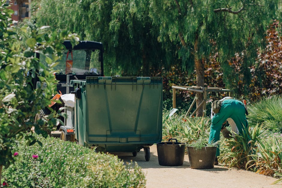 A person wearing black trousers and black sneakers is using a green rubber hand rake to gather fallen leaves, small branches, and garden debris from a gravel pathway in a garden. The rake blade is wide and textured, suitable for collecting loose material. The surrounding area includes a section of compacted soil bordered by a stone wall in the background, with some green plants growing near the base of the wall. The garden appears tidy and well-maintained, with a mix of leaf litter and small twigs on the ground, indicative of ongoing garden maintenance or hedge trimming activities. The lighting suggests it is daytime, with natural light illuminating the scene, emphasizing the textures of the gravel, leaves, and stone wall. This image relates to outdoor garden care, demonstrating the process of clearing debris as part of lawn or hedge trimming, an activity supported by Gardeners Bayswater in line with Westminster Council garden waste rules for Bayswater gardeners.